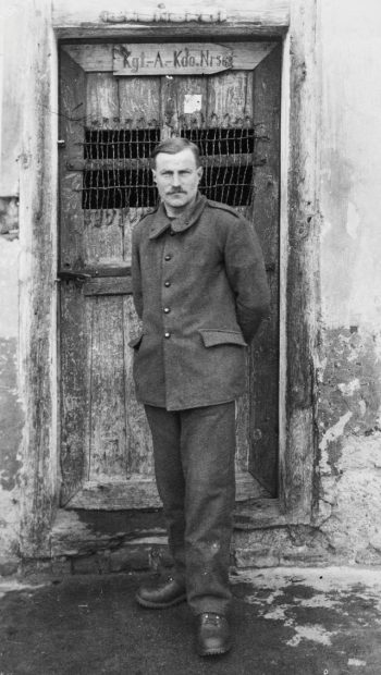 Foto von Alphonse Courtin, Stalag VII A, Gefangenennummer 22675 Courtin posiert in auffallend guter Kleidung vor der vorschriftsmäßig beschrifteten und vergitterten Tür seiner Unterkunft während seines Arbeitseinsatzes.